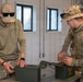 Two military personnel in multicam camouflage uniforms are working with olive drab ammunition cans on a table inside a building. A third individual is partially visible in the background. The setting appears to be a military facility or range building based on the environment and equipment present.