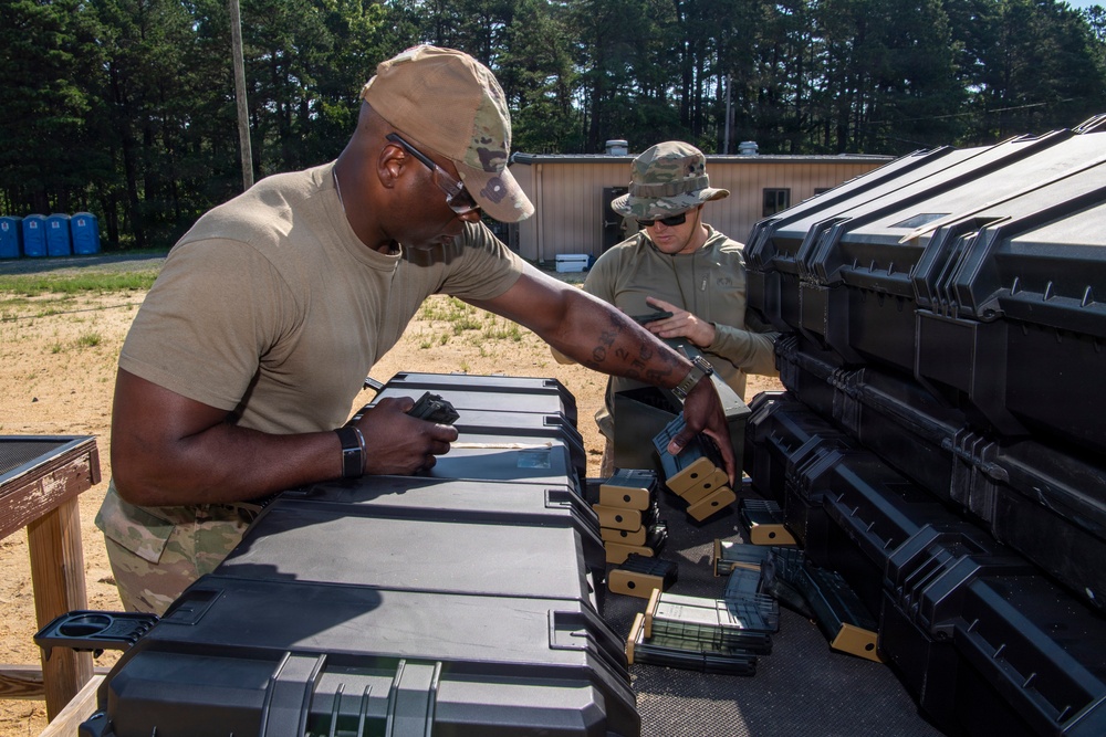 Two military personnel in OCP (Operational Camouflage Pattern) uniforms are handling equipment at what appears to be an outdoor military range or staging area. They are working with black hard-shell cases and what appear to be tan/green ammunition magazines or similar military equipment. The setting includes a sandy terrain with trees and a small building visible in the background.
