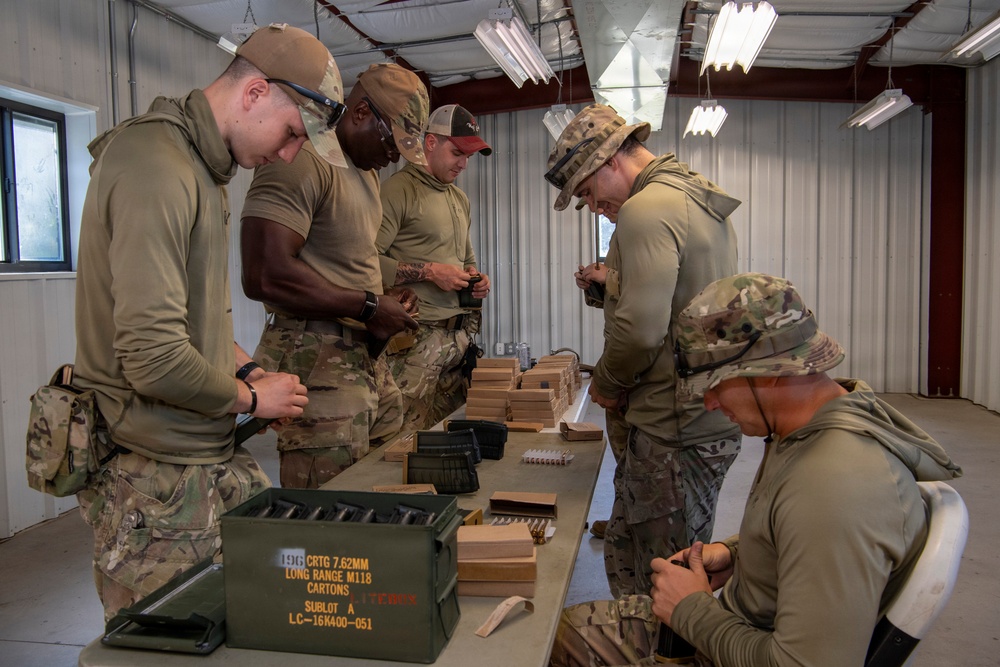 Five military personnel in OCP/MultiCam uniforms are gathered around a table inside a metal building, loading ammunition magazines. An open green ammunition can labeled '196 CRTG 7.62MM LONG RANGE M118 CARTONS SUBLOT A LC-16K400-051' is visible in the foreground. The soldiers appear to be conducting pre-range preparation by loading 7.62mm rounds into magazines.