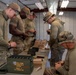 Five military personnel in OCP/MultiCam uniforms are gathered around a table inside a metal building, loading ammunition magazines. An open green ammunition can labeled '196 CRTG 7.62MM LONG RANGE M118 CARTONS SUBLOT A LC-16K400-051' is visible in the foreground. The soldiers appear to be conducting pre-range preparation by loading 7.62mm rounds into magazines.