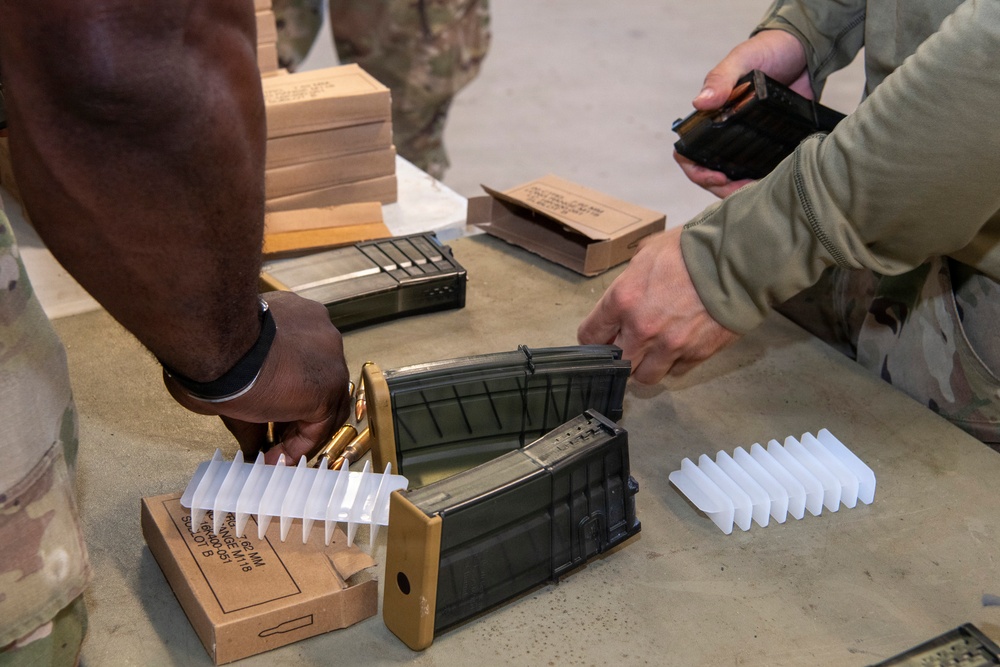 Military personnel in camouflage uniforms are loading ammunition magazines at a table. The scene shows a ammunition preparation station with multiple rifle magazines, loose rounds, and ammunition packaging boxes. Two individuals are actively handling magazines and cartridges, suggesting pre-range or pre-mission ammunition loading.