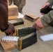 Military personnel in camouflage uniforms are loading ammunition magazines at a table. The scene shows a ammunition preparation station with multiple rifle magazines, loose rounds, and ammunition packaging boxes. Two individuals are actively handling magazines and cartridges, suggesting pre-range or pre-mission ammunition loading.