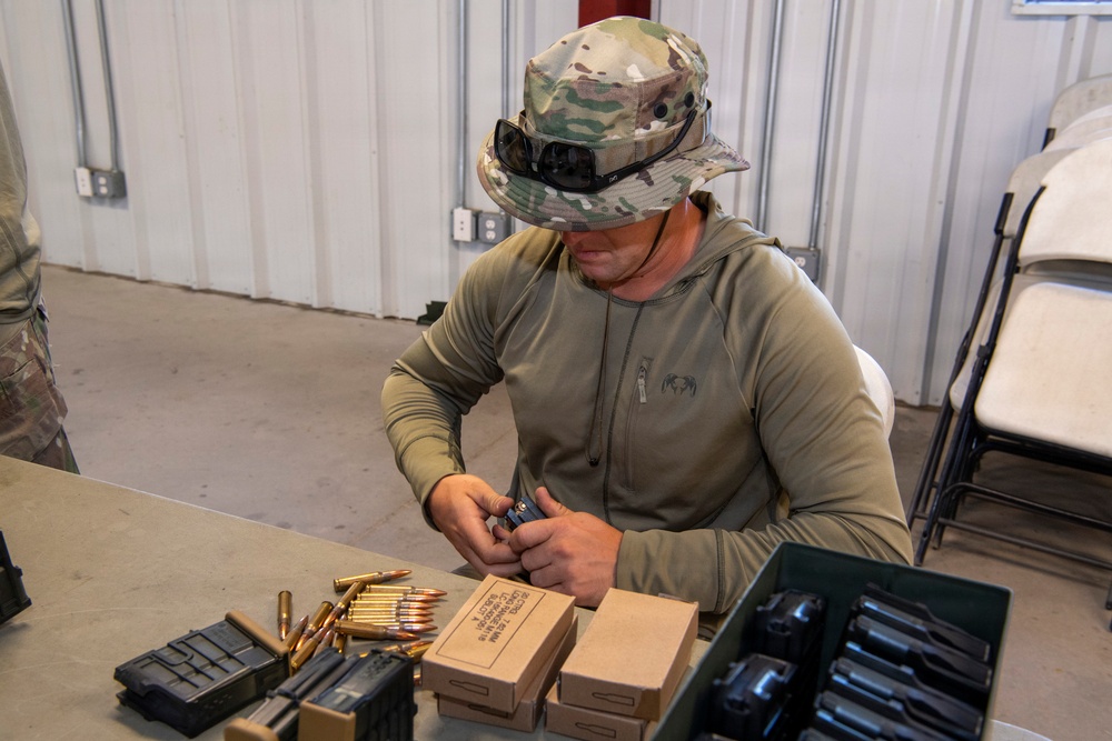 A person wearing multicam camouflage boonie hat and olive drab hoodie is seated at a table loading ammunition into a magazine. The setting appears to be an indoor range or staging area with metal wall paneling. Multiple magazines, loose rifle cartridges, and ammunition boxes are visible on the table.
