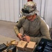 A person wearing multicam camouflage boonie hat and olive drab hoodie is seated at a table loading ammunition into a magazine. The setting appears to be an indoor range or staging area with metal wall paneling. Multiple magazines, loose rifle cartridges, and ammunition boxes are visible on the table.