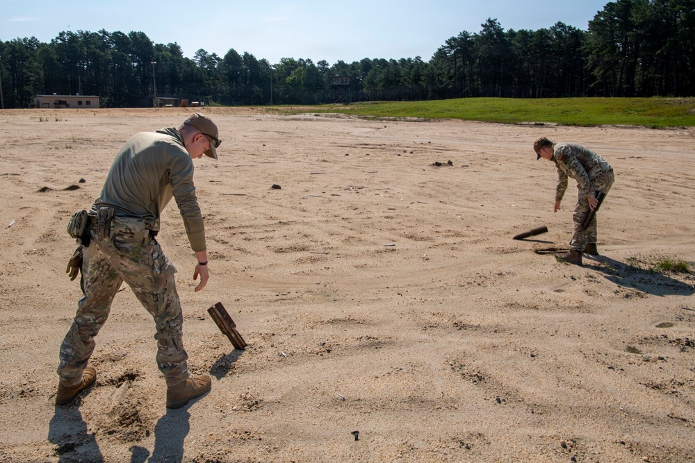 Two military personnel in camouflage uniforms are examining or collecting objects from a sandy open field or training area. Both individuals are bent over, inspecting dark cylindrical or rectangular objects partially embedded in the sand. The setting appears to be a military training range or cleared field, with tree lines and structures visible in the background.