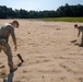 Two military personnel in camouflage uniforms are examining or collecting objects from a sandy open field or training area. Both individuals are bent over, inspecting dark cylindrical or rectangular objects partially embedded in the sand. The setting appears to be a military training range or cleared field, with tree lines and structures visible in the background.