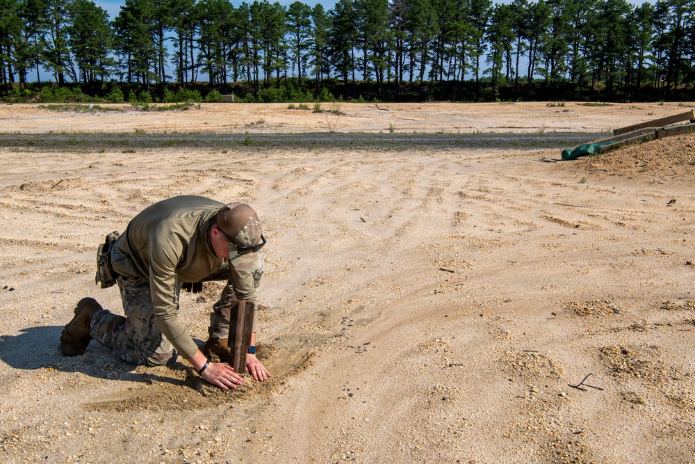 A person in military-style olive drab clothing and tactical gear is kneeling on sandy ground, working with what appears to be a wooden object partially buried or being placed in the sand. The setting appears to be a large open sandy training area or range, bordered by a tree line in the background. The activity suggests military or law enforcement training, possibly related to explosive ordnance disposal or field engineering exercises.