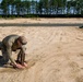 A person in military-style olive drab clothing and tactical gear is kneeling on sandy ground, working with what appears to be a wooden object partially buried or being placed in the sand. The setting appears to be a large open sandy training area or range, bordered by a tree line in the background. The activity suggests military or law enforcement training, possibly related to explosive ordnance disposal or field engineering exercises.