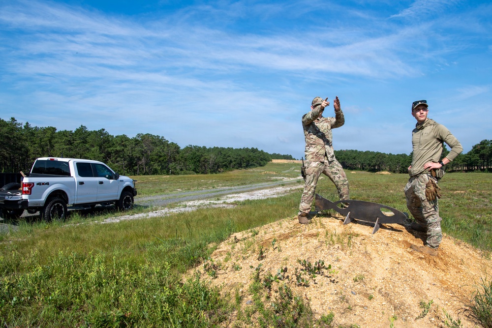 Two individuals in military/tactical camouflage uniforms are standing on a dirt berm at what appears to be an outdoor shooting range or training area. One person is gesturing with raised hands while the other stands observing, with a metal silhouette target visible between them. A white Ford F-150 4x4 pickup truck is parked nearby on a gravel path.