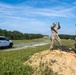 Two individuals in military/tactical camouflage uniforms are standing on a dirt berm at what appears to be an outdoor shooting range or training area. One person is gesturing with raised hands while the other stands observing, with a metal silhouette target visible between them. A white Ford F-150 4x4 pickup truck is parked nearby on a gravel path.