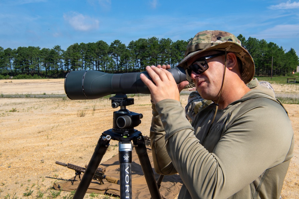 A man in military-style clothing and a camouflage boonie hat is using a large spotting scope mounted on a Vortex tripod at what appears to be an outdoor shooting range. A firearm (rifle) is visible on the ground behind him, and another person in similar attire is partially visible in the background. The setting is a sandy, open range area with trees in the background under a clear blue sky.