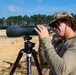 A man in military-style clothing and a camouflage boonie hat is using a large spotting scope mounted on a Vortex tripod at what appears to be an outdoor shooting range. A firearm (rifle) is visible on the ground behind him, and another person in similar attire is partially visible in the background. The setting is a sandy, open range area with trees in the background under a clear blue sky.