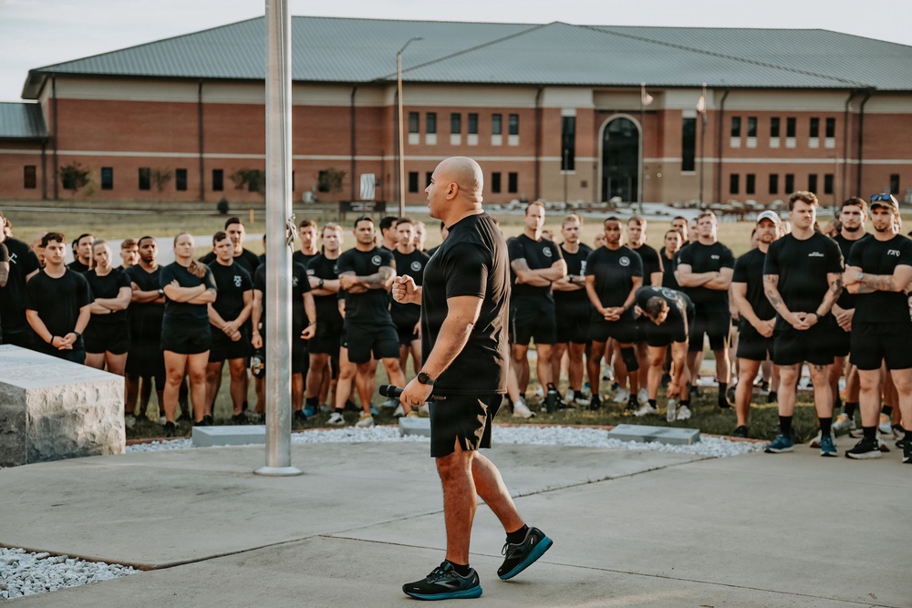 A bald man in black athletic wear appears to be addressing a large group of individuals, all uniformly dressed in black t-shirts and shorts, standing in formation outdoors. The setting appears to be a training or institutional facility with a large brick building visible in the background. The scene suggests a formal briefing, orientation, or motivational address, possibly at a law enforcement, military, or athletic training facility.