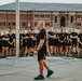 A bald man in black athletic wear appears to be addressing a large group of individuals, all uniformly dressed in black t-shirts and shorts, standing in formation outdoors. The setting appears to be a training or institutional facility with a large brick building visible in the background. The scene suggests a formal briefing, orientation, or motivational address, possibly at a law enforcement, military, or athletic training facility.