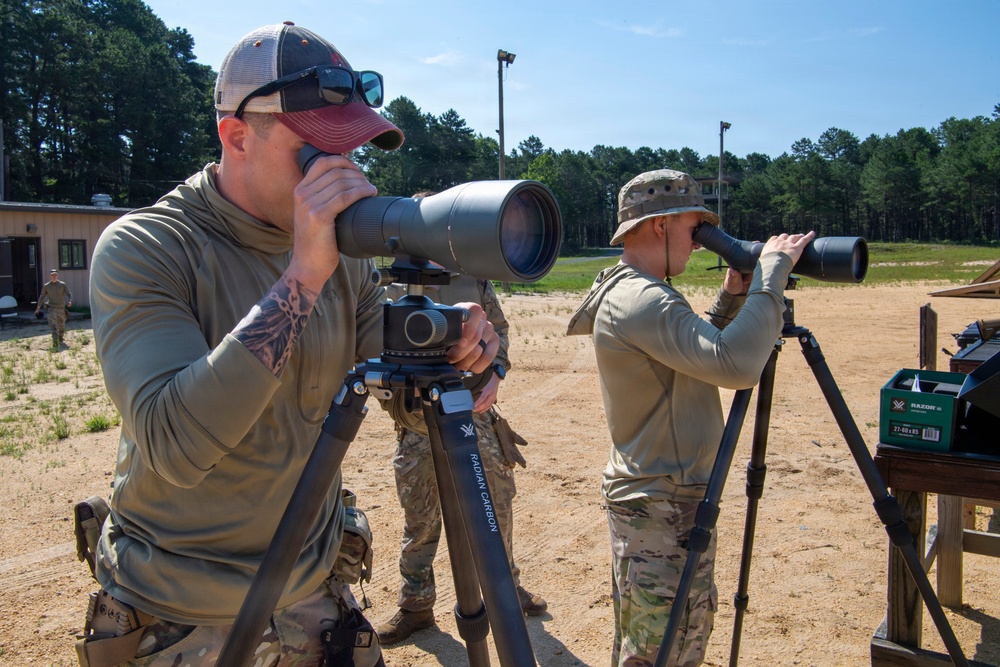Two individuals in military-style camouflage clothing are using spotting scopes mounted on tripods at what appears to be an outdoor shooting range. The foreground figure wears a baseball cap with sunglasses and has visible tattoos, while the background figure wears a boonie hat. A third person in uniform is visible in the far background near a building.