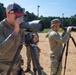 Two individuals in military-style camouflage clothing are using spotting scopes mounted on tripods at what appears to be an outdoor shooting range. The foreground figure wears a baseball cap with sunglasses and has visible tattoos, while the background figure wears a boonie hat. A third person in uniform is visible in the far background near a building.
