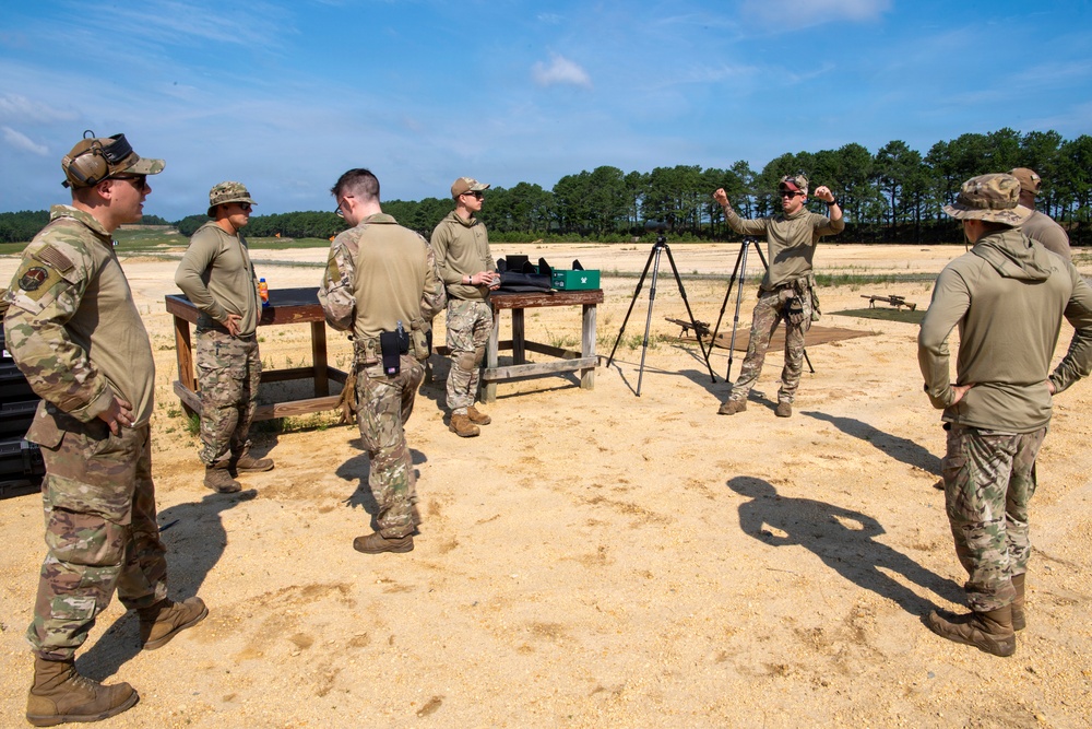 A group of military personnel in camouflage uniforms are gathered at an outdoor shooting range during a training session. One individual appears to be an instructor gesturing and briefing the group near a tripod-mounted spotting scope and a wooden table with equipment. The setting is a sandy open range area with tree lines visible in the background.
