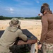 Two individuals are at an outdoor shooting range, with one person in a prone-adjacent position aiming a firearm downrange while the other stands nearby observing and gesturing. The setting appears to be a military or law enforcement training facility with a wide sandy range and tree line in the background. Both individuals are wearing tactical or military-style clothing.