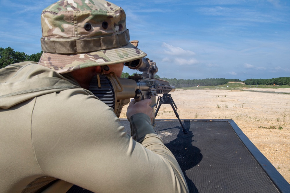 A person in military-style camouflage clothing and a boonie hat is in a prone shooting position on a firing range, aiming a precision rifle equipped with a bipod and optical scope. The setting appears to be an outdoor military or law enforcement shooting range with a long sandy downrange area and tree line in the background. The image is taken from behind and slightly to the side of the shooter.