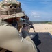 A person in military-style camouflage clothing and a boonie hat is in a prone shooting position on a firing range, aiming a precision rifle equipped with a bipod and optical scope. The setting appears to be an outdoor military or law enforcement shooting range with a long sandy downrange area and tree line in the background. The image is taken from behind and slightly to the side of the shooter.