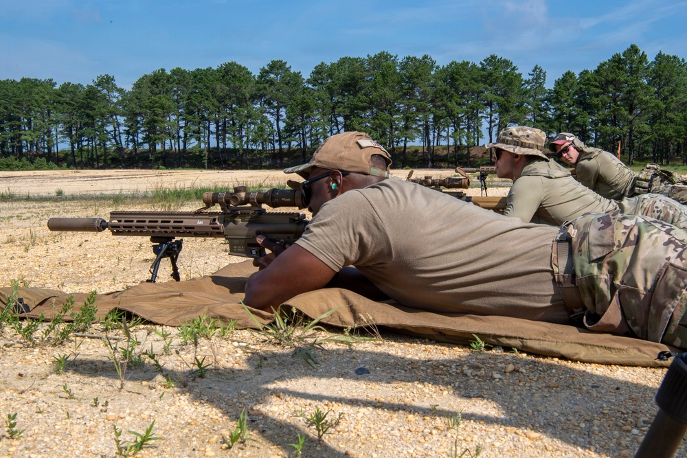 Military or law enforcement personnel are conducting a rifle marksmanship training exercise at an outdoor shooting range. A shooter in the prone position is aiming a suppressed precision rifle equipped with a bipod and optical scope, while two observers positioned nearby appear to be coaching or evaluating. The setting is a sandy open range with a tree line in the background under clear skies.