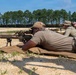 Military or law enforcement personnel are conducting a rifle marksmanship training exercise at an outdoor shooting range. A shooter in the prone position is aiming a suppressed precision rifle equipped with a bipod and optical scope, while two observers positioned nearby appear to be coaching or evaluating. The setting is a sandy open range with a tree line in the background under clear skies.