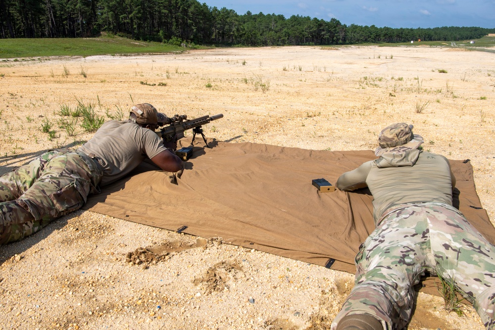 Two military personnel in camouflage uniforms are lying prone on a tan shooting mat at an outdoor firing range. The shooter on the left is aiming a rifle equipped with a bipod and suppressor downrange, while the second individual on the right appears to be observing or spotting. The setting is a sandy open range with a tree line visible in the background.