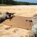 Two military personnel in camouflage uniforms are lying prone on a tan shooting mat at an outdoor firing range. The shooter on the left is aiming a rifle equipped with a bipod and suppressor downrange, while the second individual on the right appears to be observing or spotting. The setting is a sandy open range with a tree line visible in the background.
