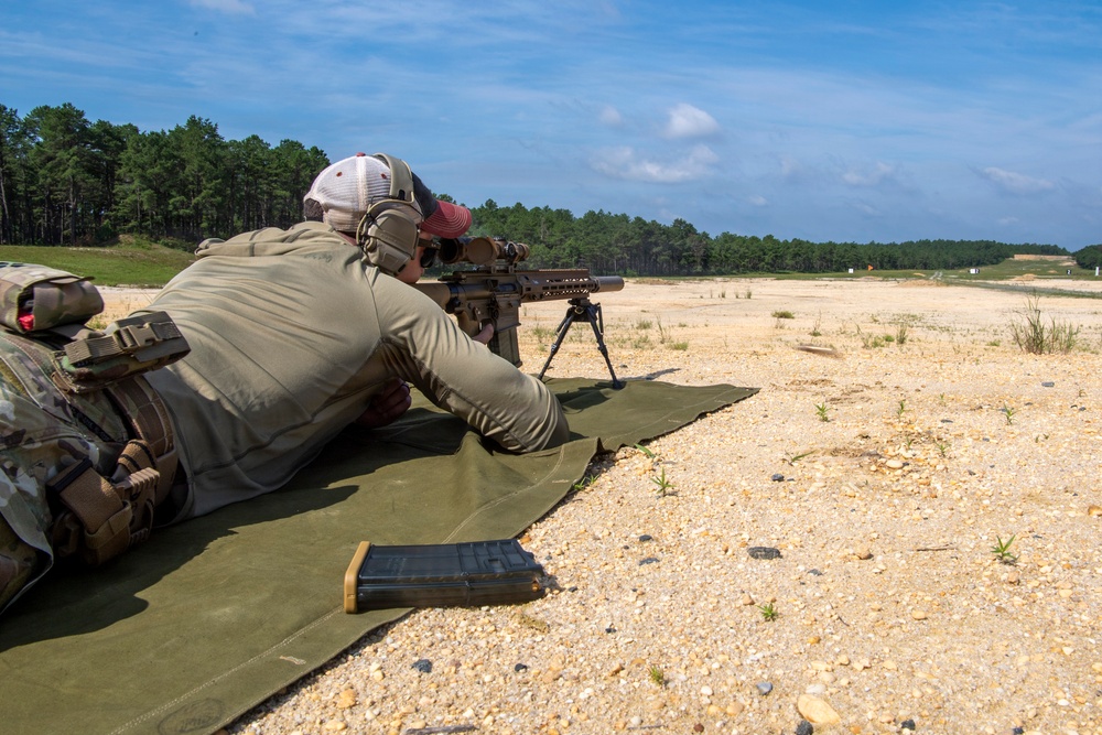 A shooter is in a prone position on a green mat at an outdoor shooting range, aiming a scoped rifle mounted on a bipod downrange. The setting appears to be a military or law enforcement training facility based on the equipment and range layout. Sandy terrain, distant target markers, and a treeline are visible in the background.