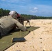 A shooter is in a prone position on a green mat at an outdoor shooting range, aiming a scoped rifle mounted on a bipod downrange. The setting appears to be a military or law enforcement training facility based on the equipment and range layout. Sandy terrain, distant target markers, and a treeline are visible in the background.