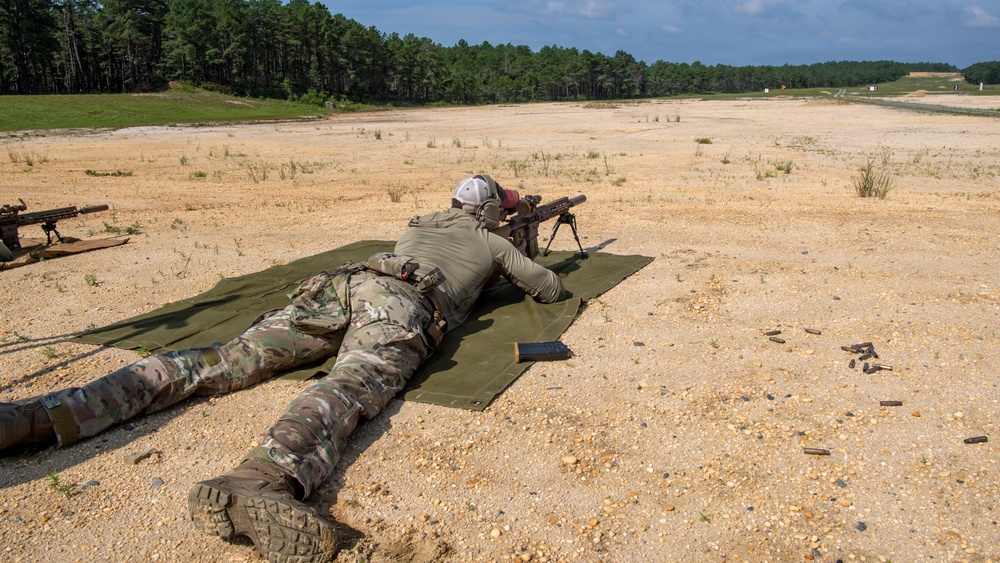 A shooter in military camouflage uniform is in a prone position on a green mat at an outdoor firing range, aiming a rifle mounted on a bipod downrange. Spent brass casings are visible on the ground to the right, indicating active firing has occurred. A second rifle on a bipod is visible in the background to the left.