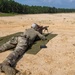 A shooter in military camouflage uniform is in a prone position on a green mat at an outdoor firing range, aiming a rifle mounted on a bipod downrange. Spent brass casings are visible on the ground to the right, indicating active firing has occurred. A second rifle on a bipod is visible in the background to the left.