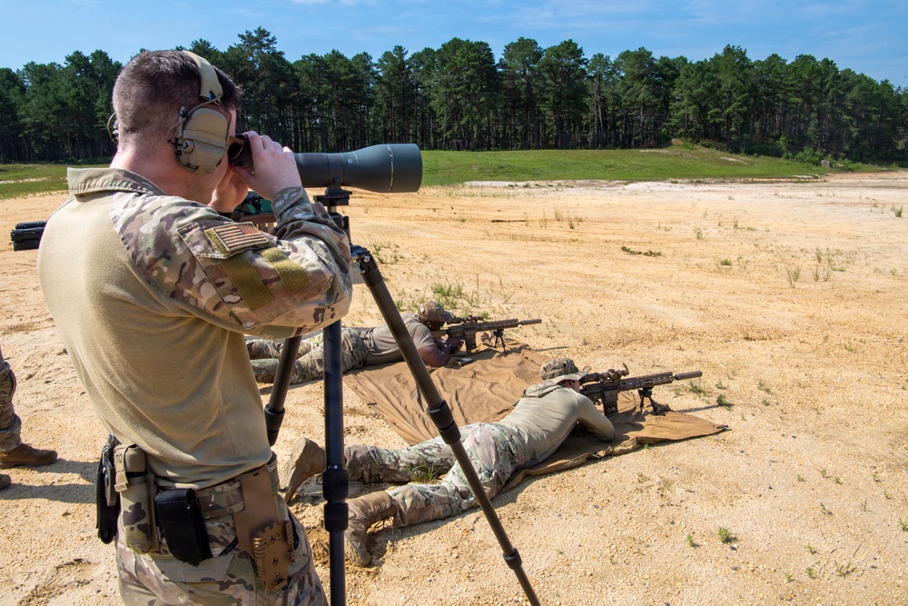 Military personnel are conducting a live-fire sniper training exercise at an outdoor shooting range. A standing soldier acts as a spotter using a large spotting scope on a tripod, while at least two other soldiers are in the prone position firing rifles downrange. The setting appears to be a sandy, open range with a tree line in the background.