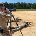 Military personnel are conducting a live-fire sniper training exercise at an outdoor shooting range. A standing soldier acts as a spotter using a large spotting scope on a tripod, while at least two other soldiers are in the prone position firing rifles downrange. The setting appears to be a sandy, open range with a tree line in the background.
