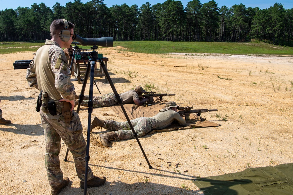 Military personnel are conducting a long-range rifle shooting exercise at an outdoor firing range. One standing soldier is observing through a spotting scope on a tripod while two others are in the prone position firing suppressed rifles downrange. The setting appears to be a sandy training range bordered by a tree line.