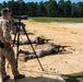 Military personnel are conducting a long-range rifle shooting exercise at an outdoor firing range. One standing soldier is observing through a spotting scope on a tripod while two others are in the prone position firing suppressed rifles downrange. The setting appears to be a sandy training range bordered by a tree line.