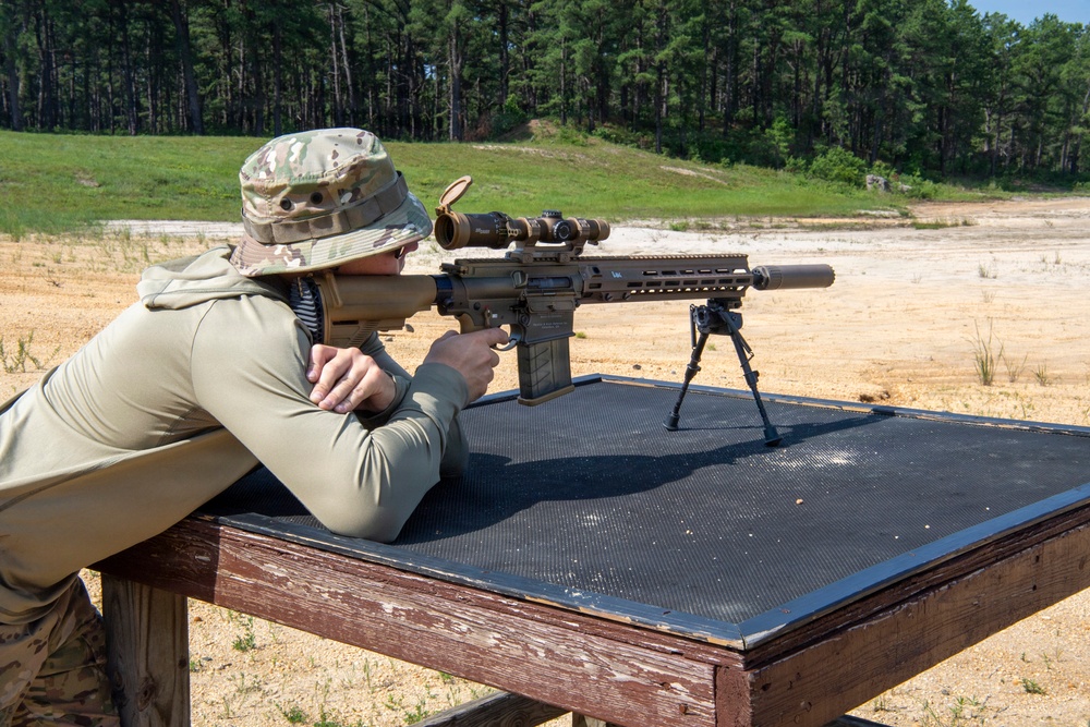 A shooter in military-style camouflage clothing is in a prone-adjacent position at a shooting bench, aiming a precision rifle equipped with a bipod, suppressor, and magnified optic. The setting appears to be an outdoor shooting range with a sandy berm and tree line in the background. The rifle is stabilized on a rubber shooting mat atop a wooden bench rest.