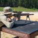 A shooter in military-style camouflage clothing is in a prone-adjacent position at a shooting bench, aiming a precision rifle equipped with a bipod, suppressor, and magnified optic. The setting appears to be an outdoor shooting range with a sandy berm and tree line in the background. The rifle is stabilized on a rubber shooting mat atop a wooden bench rest.