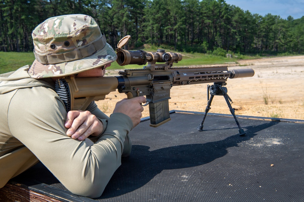A person wearing multicam camouflage clothing and a boonie hat is in a prone shooting position on a black mat at an outdoor shooting range. They are aiming a semi-automatic rifle equipped with a magnified optic scope and suppressor. A second similar rifle on a bipod is visible in the background on the same shooting bench.