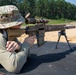 A person wearing multicam camouflage clothing and a boonie hat is in a prone shooting position on a black mat at an outdoor shooting range. They are aiming a semi-automatic rifle equipped with a magnified optic scope and suppressor. A second similar rifle on a bipod is visible in the background on the same shooting bench.