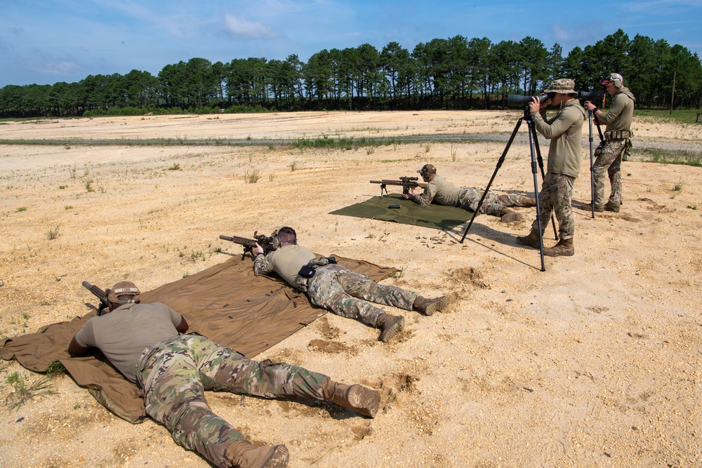 Military personnel are conducting a long-range rifle shooting exercise at an outdoor firing range. Two shooters are in prone positions on ground mats while two observers use a spotting scope on a tripod to assess downrange targets. The setting appears to be a sandy, open training range with a tree line in the background.