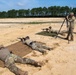 Military personnel are conducting a long-range rifle shooting exercise at an outdoor firing range. Two shooters are in prone positions on ground mats while two observers use a spotting scope on a tripod to assess downrange targets. The setting appears to be a sandy, open training range with a tree line in the background.