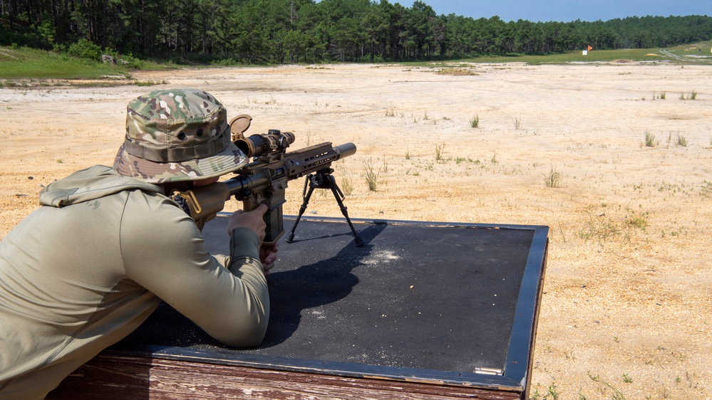 A shooter in military-style camouflage clothing and a boonie hat is in a prone position on a shooting bench, aiming a rifle equipped with a bipod, suppressor, and optical scope downrange. The setting appears to be an outdoor military or civilian shooting range with a sandy terrain and treeline in the background. A small orange marker flag is visible in the distance indicating a target area.