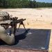A shooter in military-style camouflage clothing and a boonie hat is in a prone position on a shooting bench, aiming a rifle equipped with a bipod, suppressor, and optical scope downrange. The setting appears to be an outdoor military or civilian shooting range with a sandy terrain and treeline in the background. A small orange marker flag is visible in the distance indicating a target area.