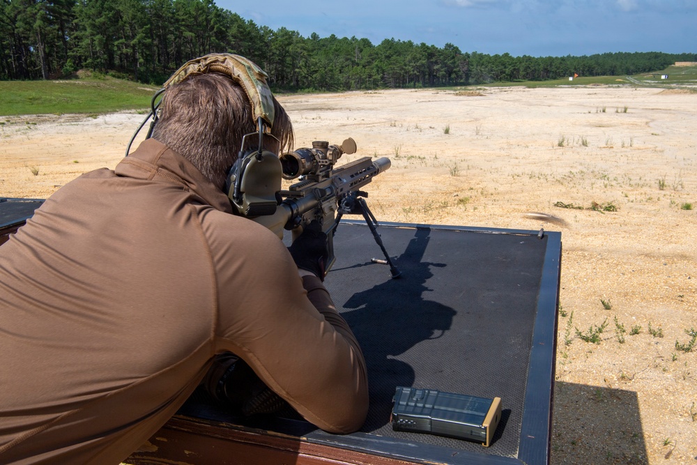 A shooter is positioned prone on a shooting bench at an outdoor firing range, aiming a scoped rifle mounted on a bipod downrange. The range appears to be a large open sandy area with tree lines visible in the background. The shooter is equipped with hearing protection and a tactical helmet.