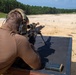 A shooter is positioned prone on a shooting bench at an outdoor firing range, aiming a scoped rifle mounted on a bipod downrange. The range appears to be a large open sandy area with tree lines visible in the background. The shooter is equipped with hearing protection and a tactical helmet.