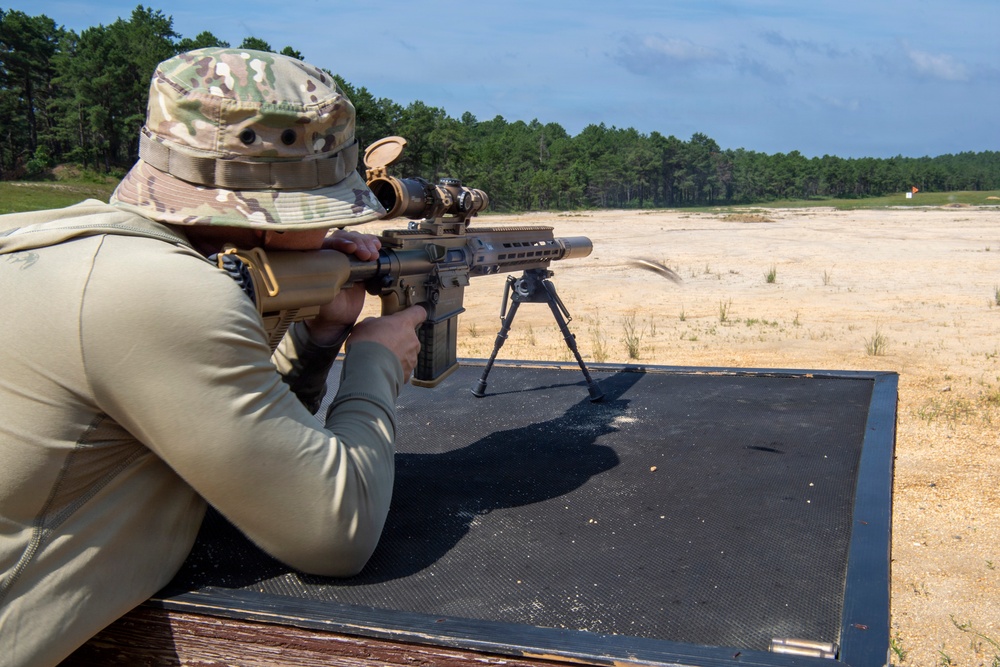 A shooter in military-style camouflage clothing and a boonie hat is in a prone position on a shooting mat, aiming a precision rifle equipped with a bipod and scope. The setting appears to be an outdoor shooting range with a sandy open field and tree line in the background. A spent casing appears to be ejecting from the rifle, suggesting the weapon has just been fired.