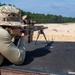 A shooter in military-style camouflage clothing and a boonie hat is in a prone position on a shooting mat, aiming a precision rifle equipped with a bipod and scope. The setting appears to be an outdoor shooting range with a sandy open field and tree line in the background. A spent casing appears to be ejecting from the rifle, suggesting the weapon has just been fired.