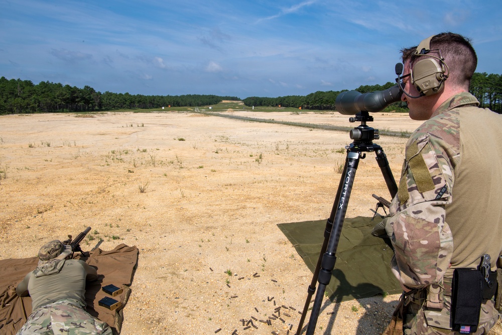 Two military personnel in MultiCam camouflage uniforms are conducting a live-fire range exercise at an outdoor shooting range. One soldier is in a prone position firing a weapon while the other acts as a spotter, using a Vortex spotting scope mounted on a tripod to observe downrange targets. Spent brass casings are visible scattered on the ground near the firing position.