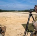 Two military personnel in MultiCam camouflage uniforms are conducting a live-fire range exercise at an outdoor shooting range. One soldier is in a prone position firing a weapon while the other acts as a spotter, using a Vortex spotting scope mounted on a tripod to observe downrange targets. Spent brass casings are visible scattered on the ground near the firing position.